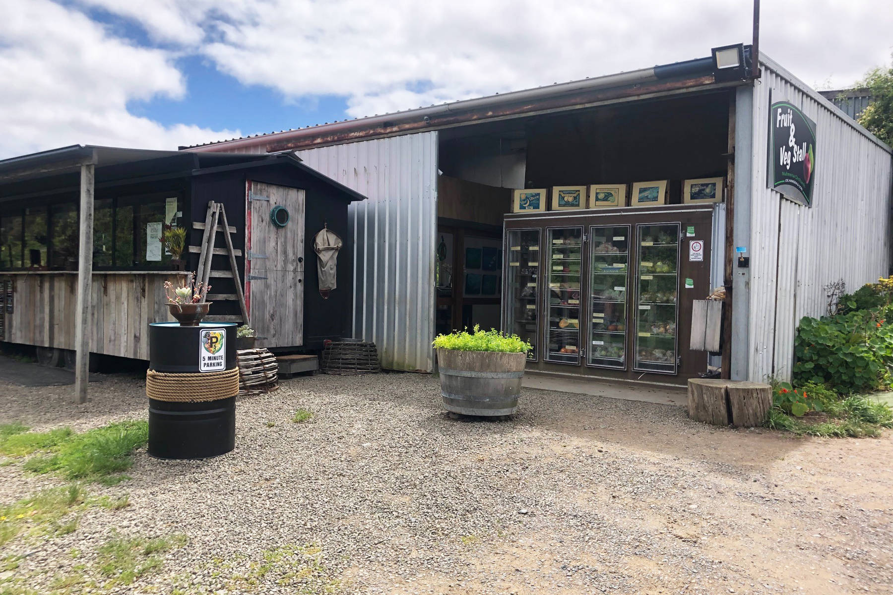 Outside view of the farmgate stall. There is a wooden building to the left of the stall and a metal barrel with a pot plant on top and a sign for 5 minute parking. A half wooden barrel filled with plants sits in front of the stall. Photo: OE Hansen.